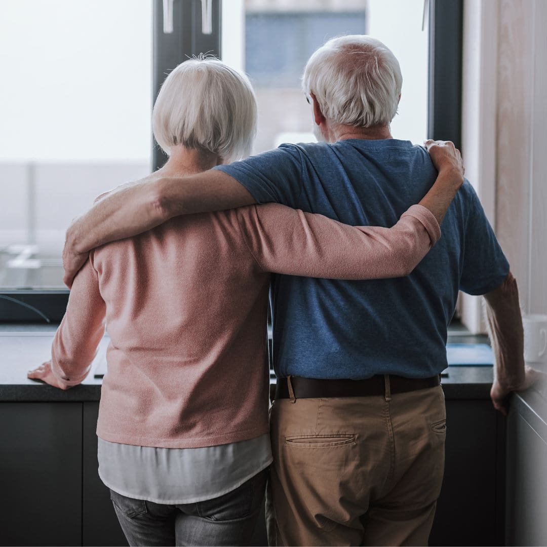 two older adults with gray hair stand side by side, arms around each other, looking out a window from inside a kitchen.