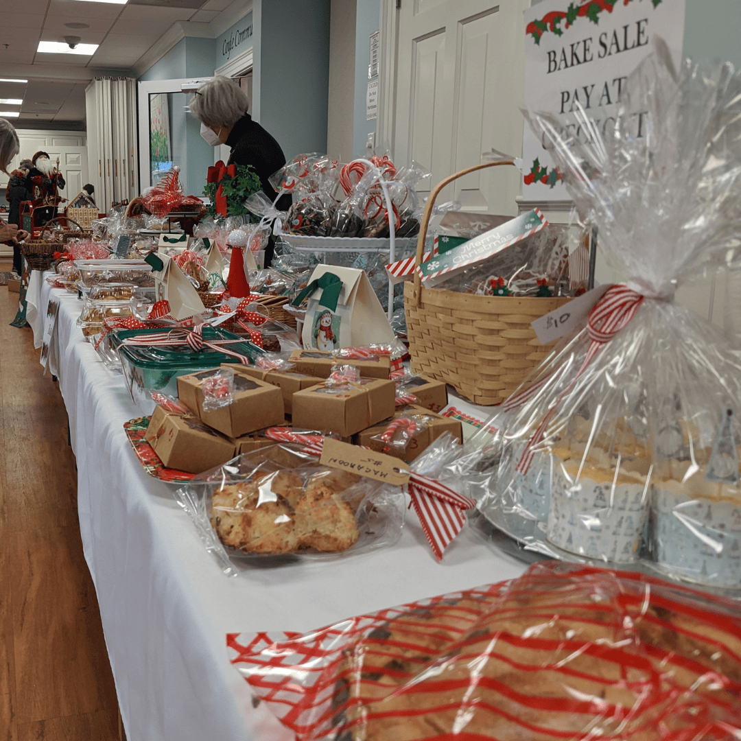 Various packaged baked goods and baskets sit on a table with a white tablecloth. A volunteer is seen in the background speaking to a shopper.