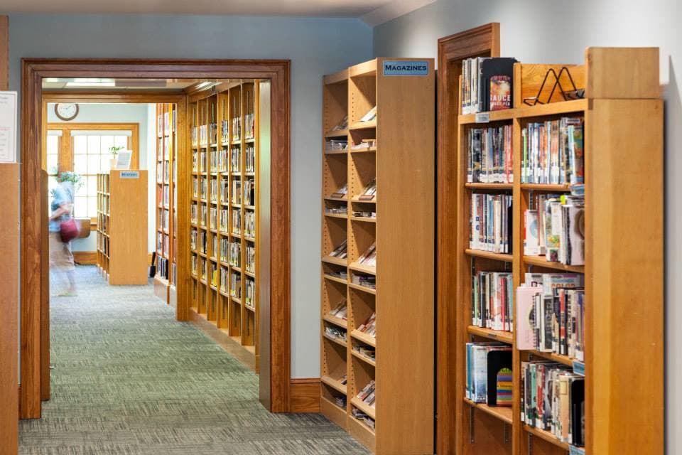 Library interior with rows of wooden bookshelves filled with books and magazines; a blurred person is walking in the background.