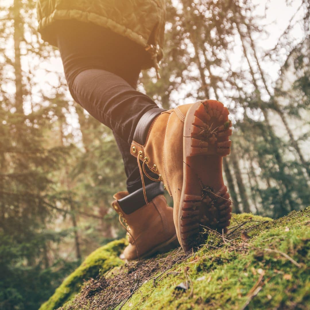Person wearing tan hiking boots walks on a mossy forest trail; photo taken from a low angle with trees in the background.
