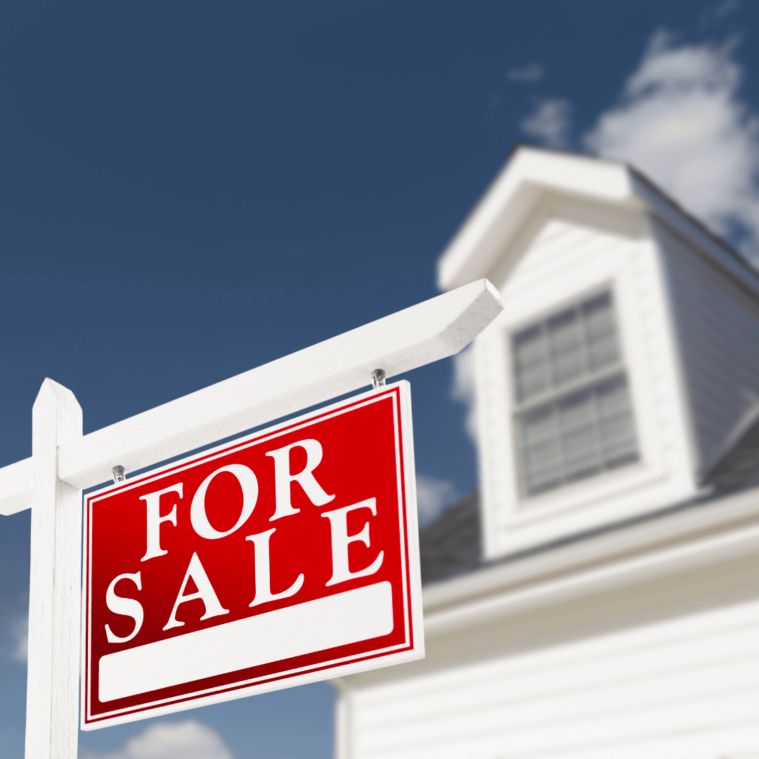 A red and white "For Sale" sign hangs in front of a white house with a gabled roof, under a blue sky with some clouds.