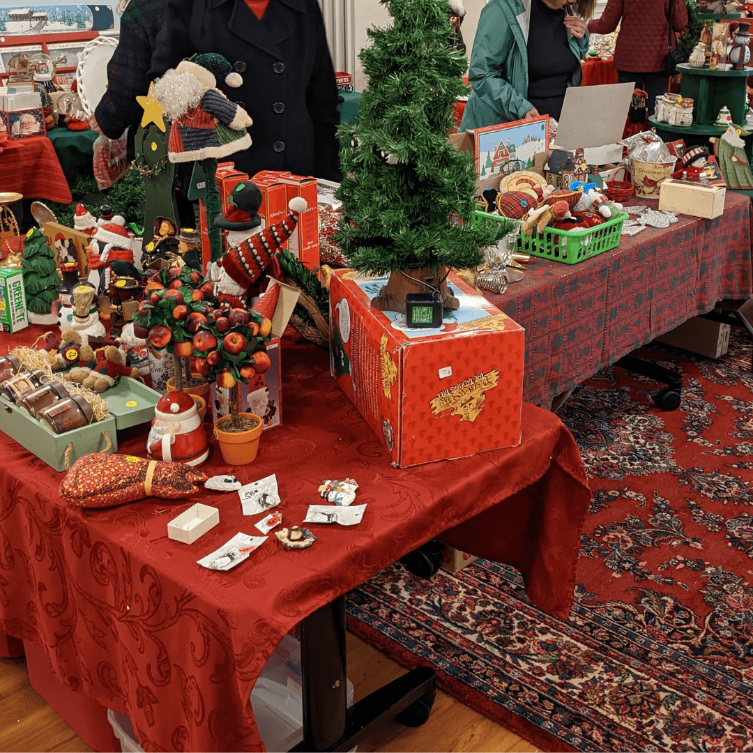 Holiday decorations are displayed on tables with red patterned tablecloths. People are seen looking at the decorations from behind the tables.