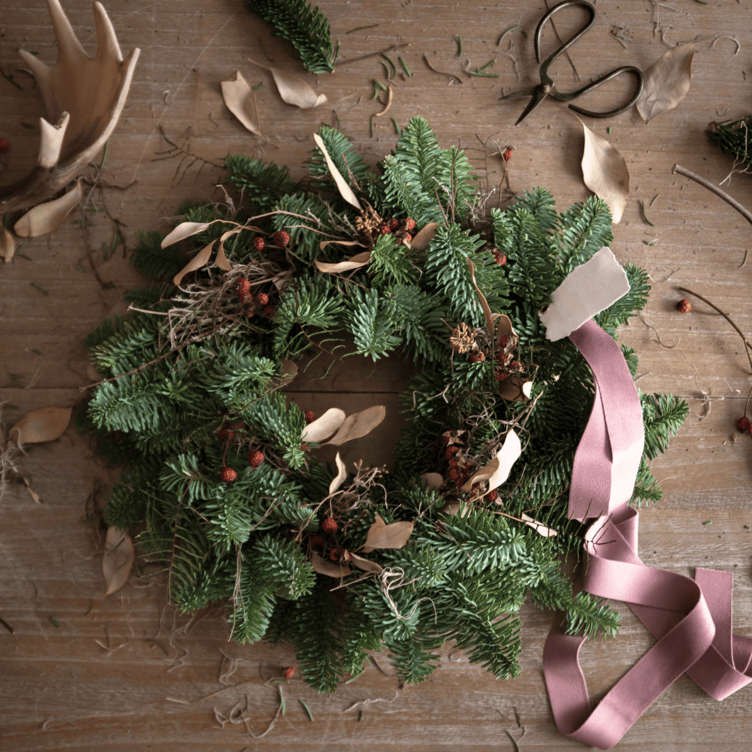 A wreath with berries and a pink ribbon sits on a wooden surface. An antler and a pair of scissors lay on either side.