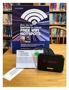 A T-Mobile WiFi hotspot device sits on a table in front of a sign advertising free WiFi hotspot borrowing at a library, with bookshelves in the background.