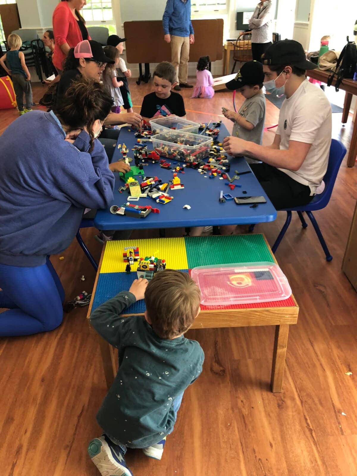 Several children and adults sit and stand around blue tables, building with LEGO bricks in a brightly lit indoor play area.