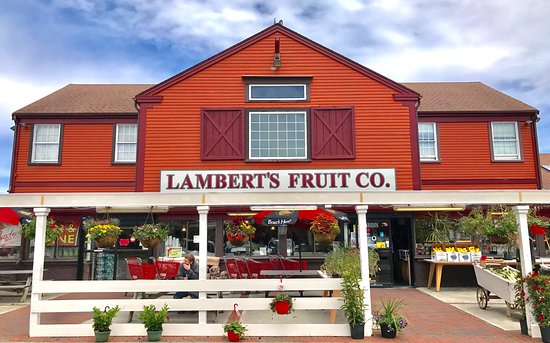 Red building with white trim labeled "Lambert's Fruit Co." displays hanging plants and produce outside under a covered porch on a brick walkway.
