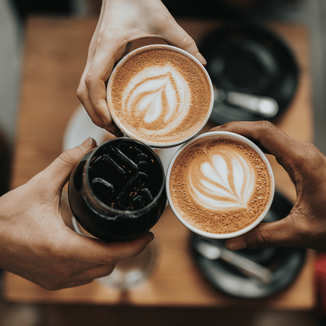 three hands holding two cups of latte with latte art and one glass of iced coffee over a wooden table with plates and cutlery in the background.