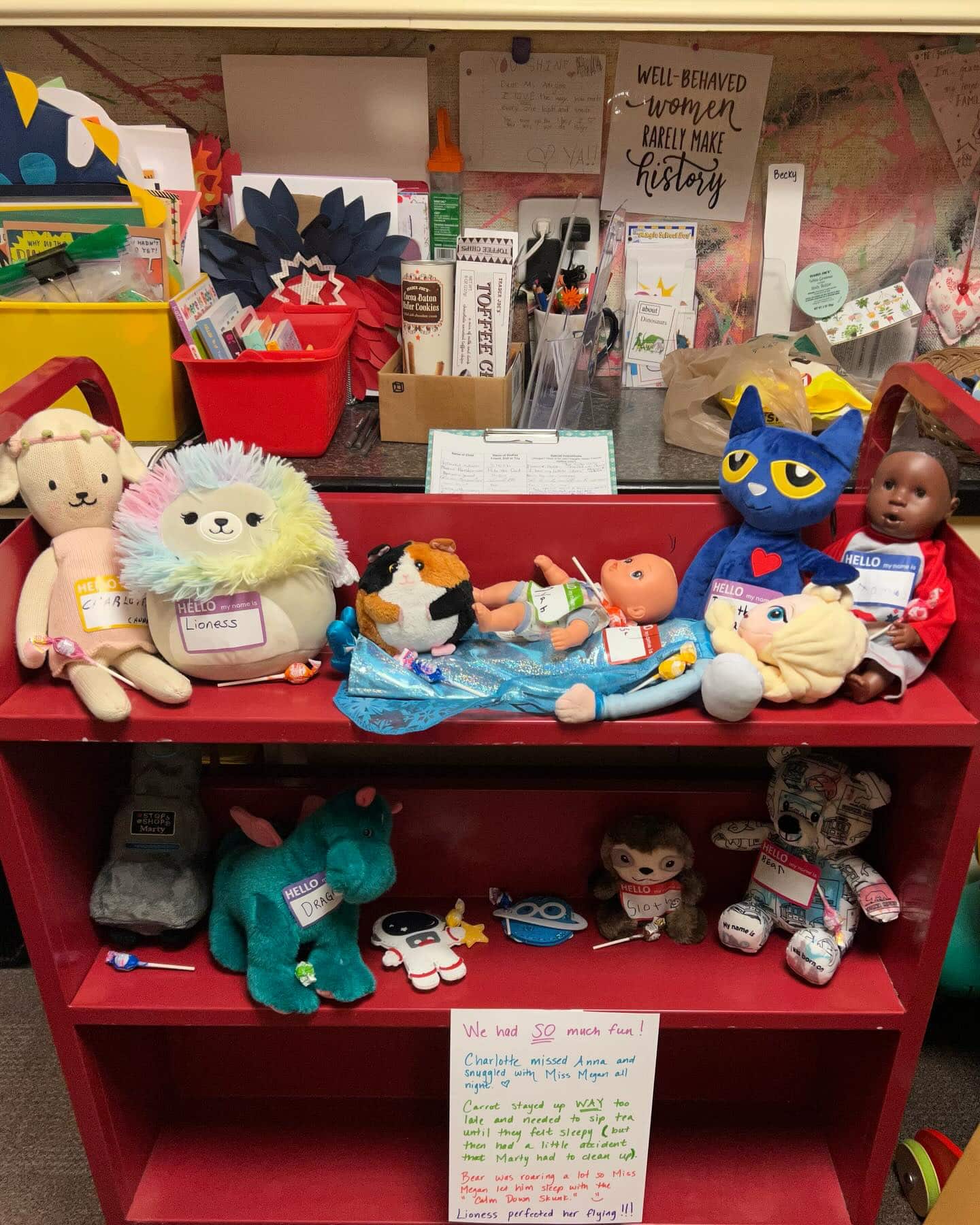 A red bookshelf displays a variety of stuffed animals and dolls, each with a name tag, in a colorful and cluttered classroom or playroom setting.