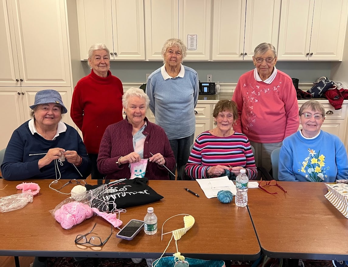 Seven elderly women sit and stand around a table with yarn, knitting needles, and water bottles, engaging in a knitting activity in a room with white cabinets.